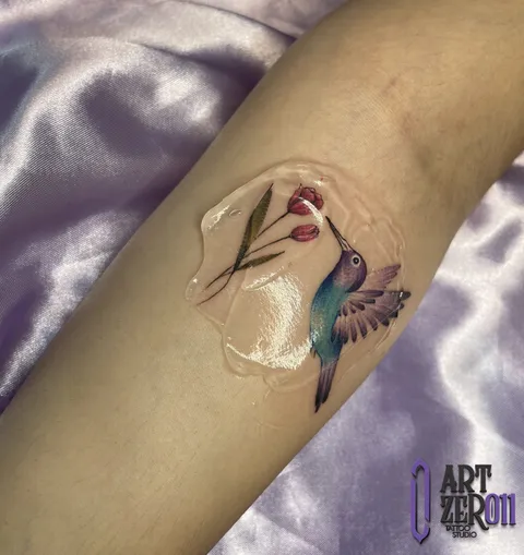 Beija-flor em aquarela azul e roxa bebendo de uma tulipa vermelha, no antebraço