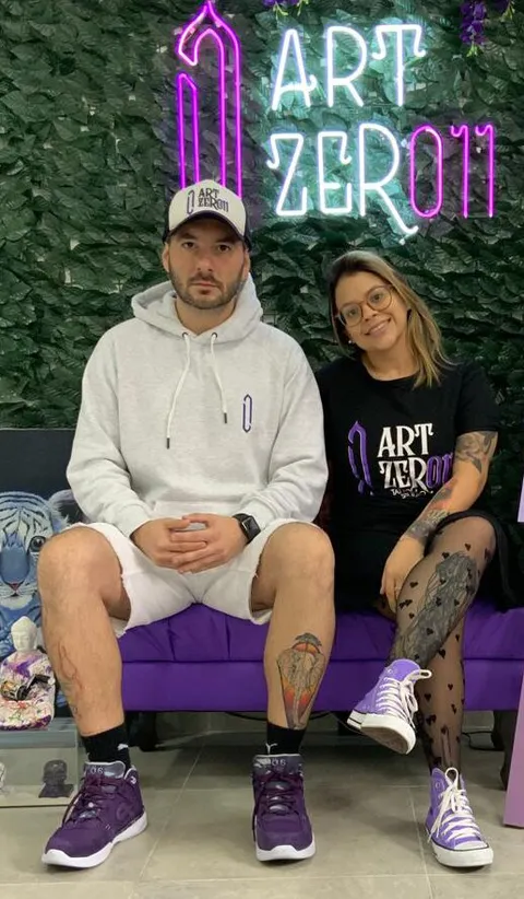 Bruno Calderelli e Flávia Borges sentados num banco roxo, em frente ao logo do ArtZero11 em neon