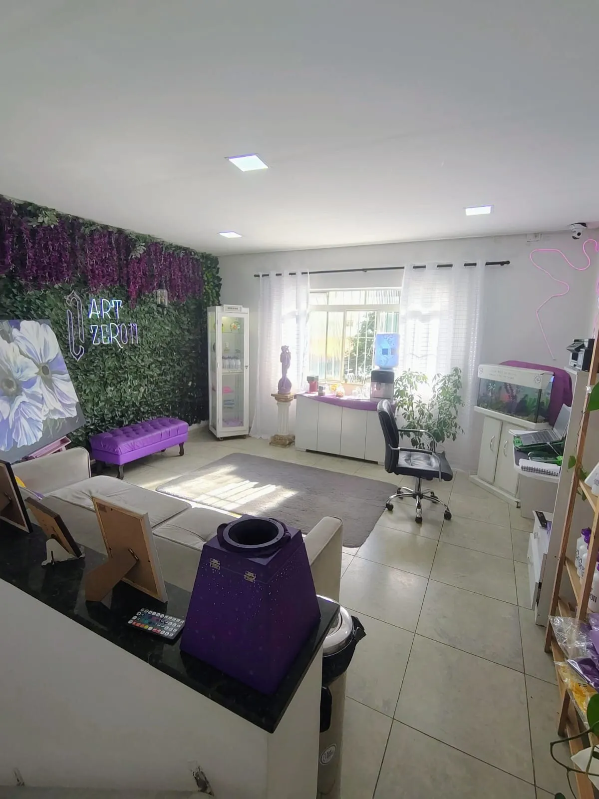 Recepção do estúdio: parede de folhagem com o logo em neon, pufe roxo, sofá e janela com luz do dia