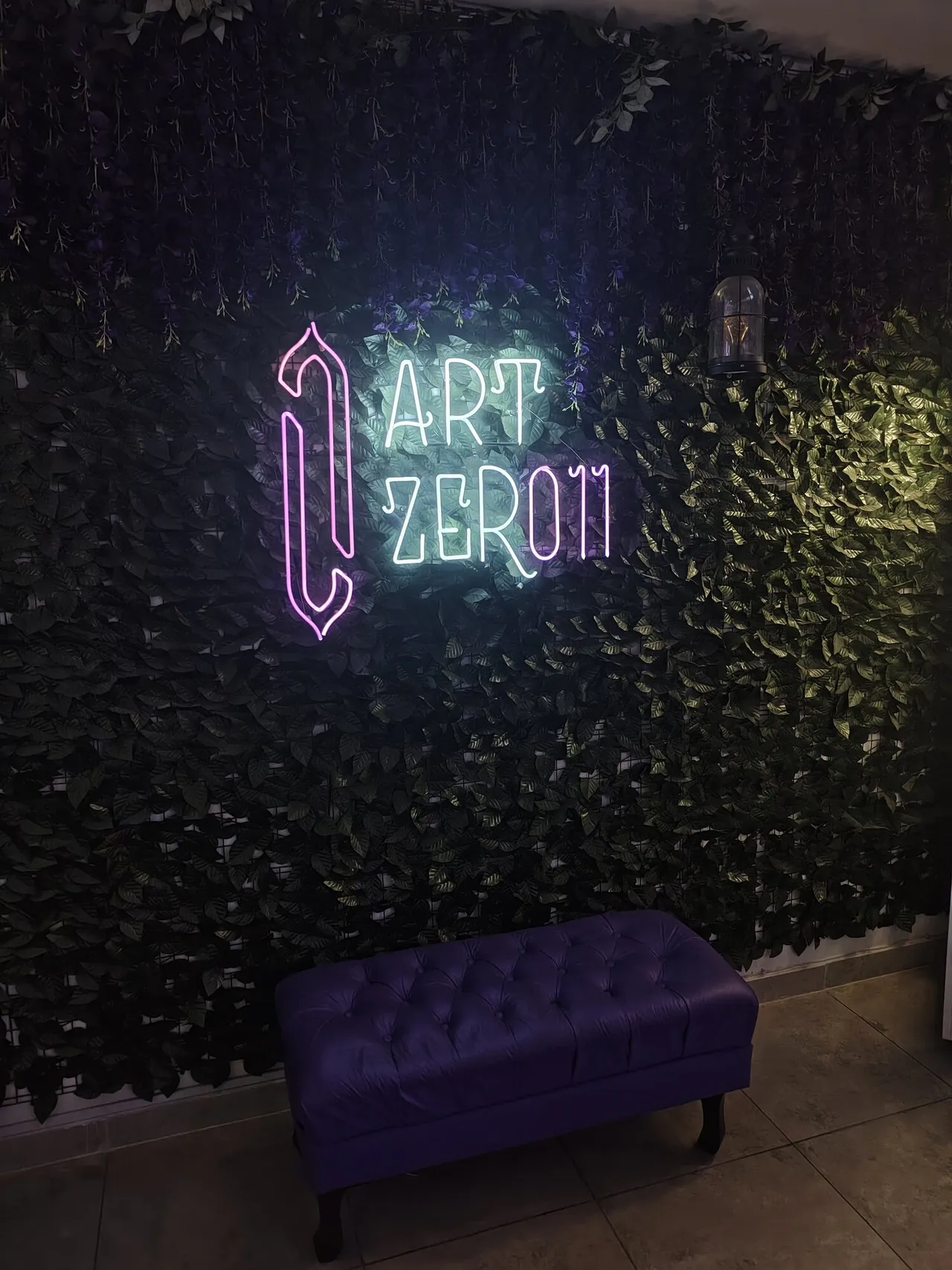 Logo do ArtZero11 em neon numa parede de folhas, com um banco roxo embaixo