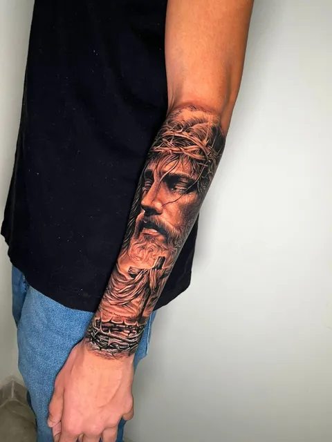 Rosto de Jesus com coroa de espinhos, em realismo preto e cinza, no antebraço